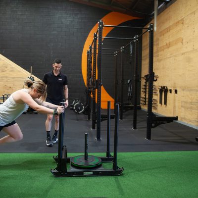Woman pushing sled under supervision of coach in o-gym on green turf